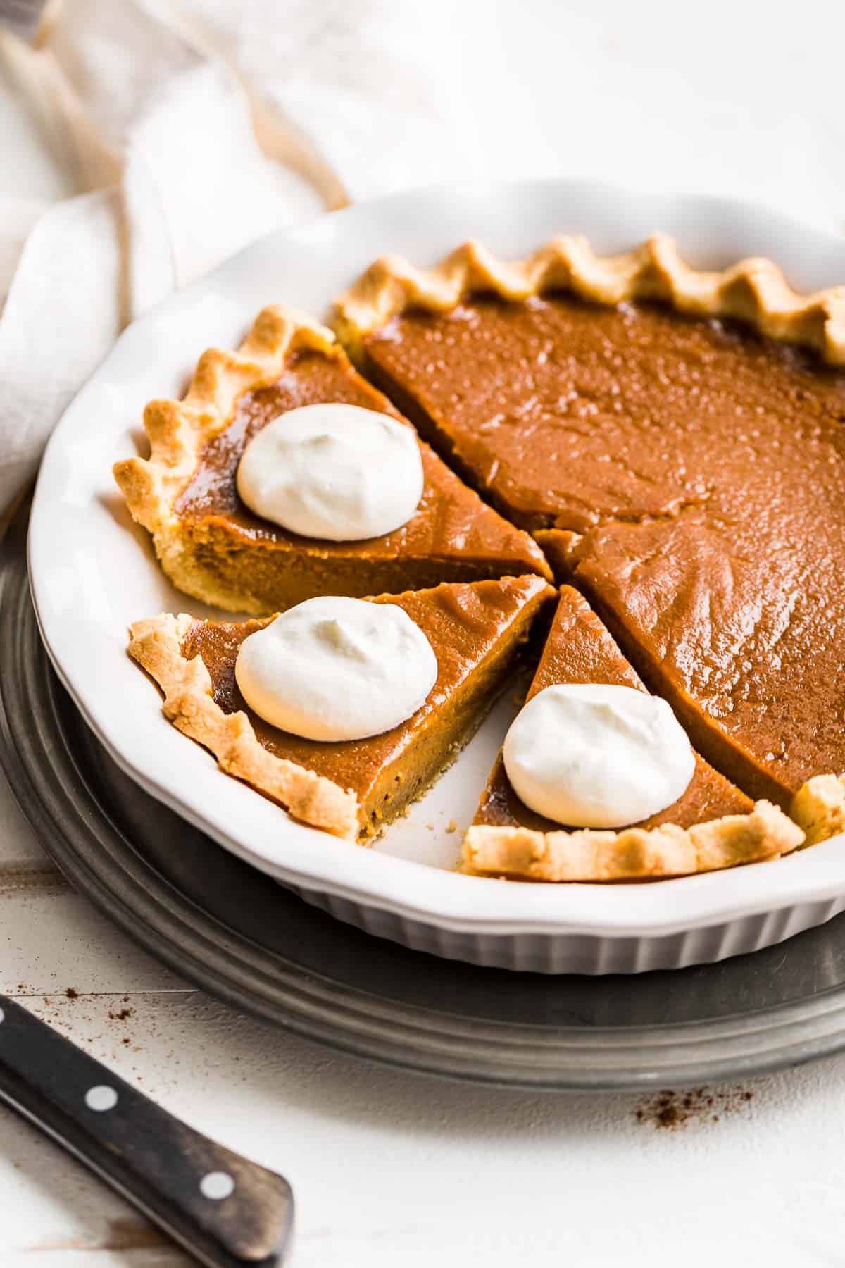 Paleo Pumpkin Pie in a white pie plate with three slices cut and topped with whipped cream dollops.