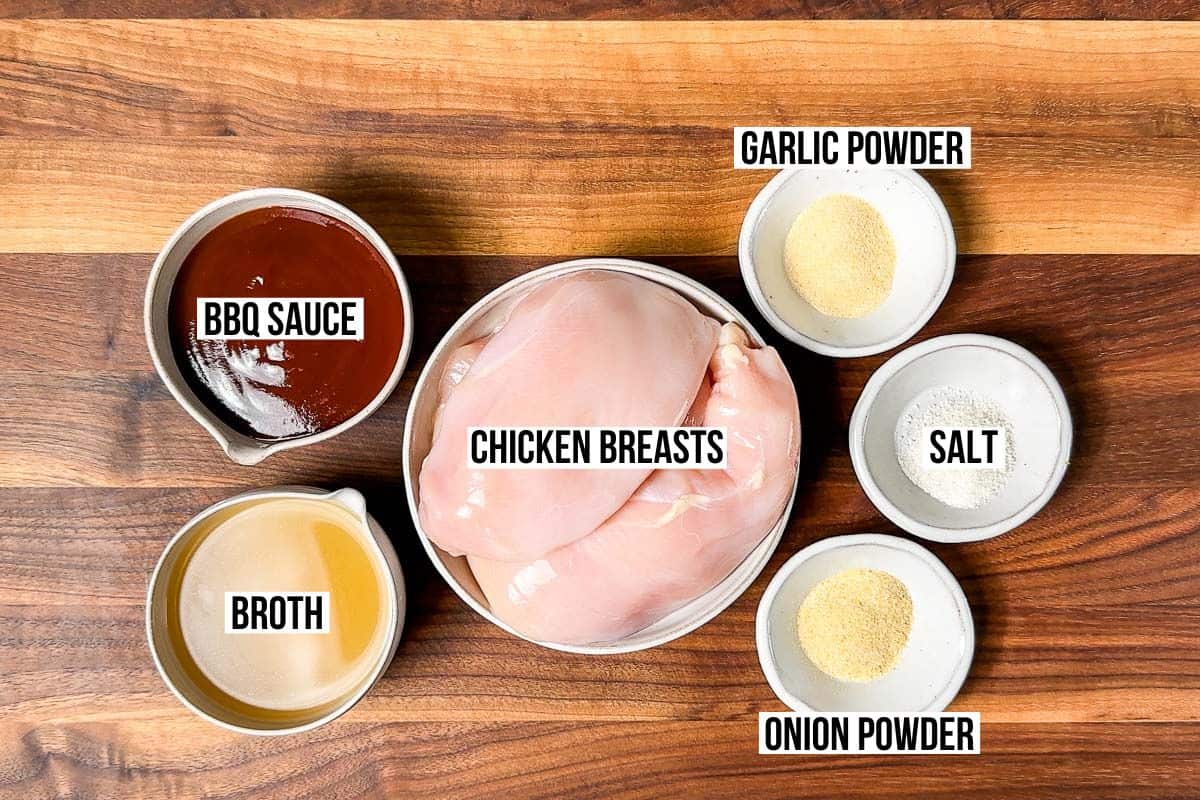 BBQ sauce, chicken broth, garlic powder, onion powder, chicken breasts and sea salt in bowls on a wood cutting board.