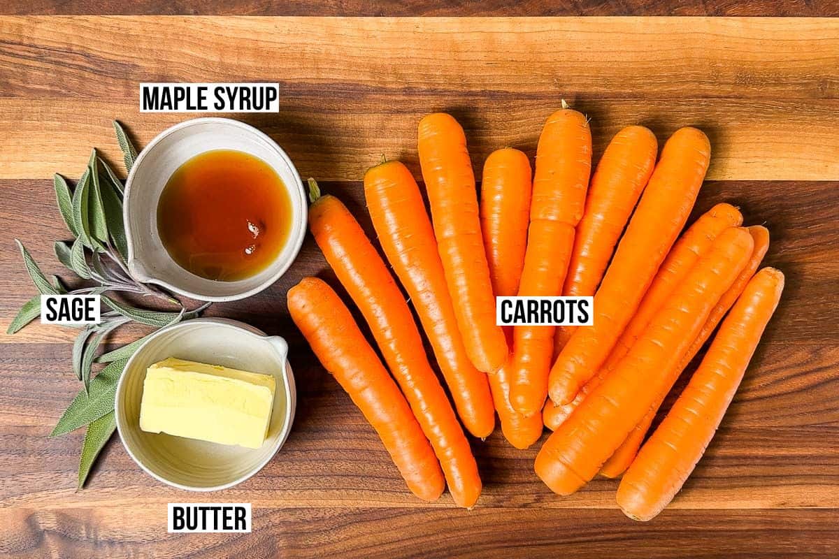 Carrots, butter, maple syrup, and sage leave in small pottery containers on a wood cutting board.