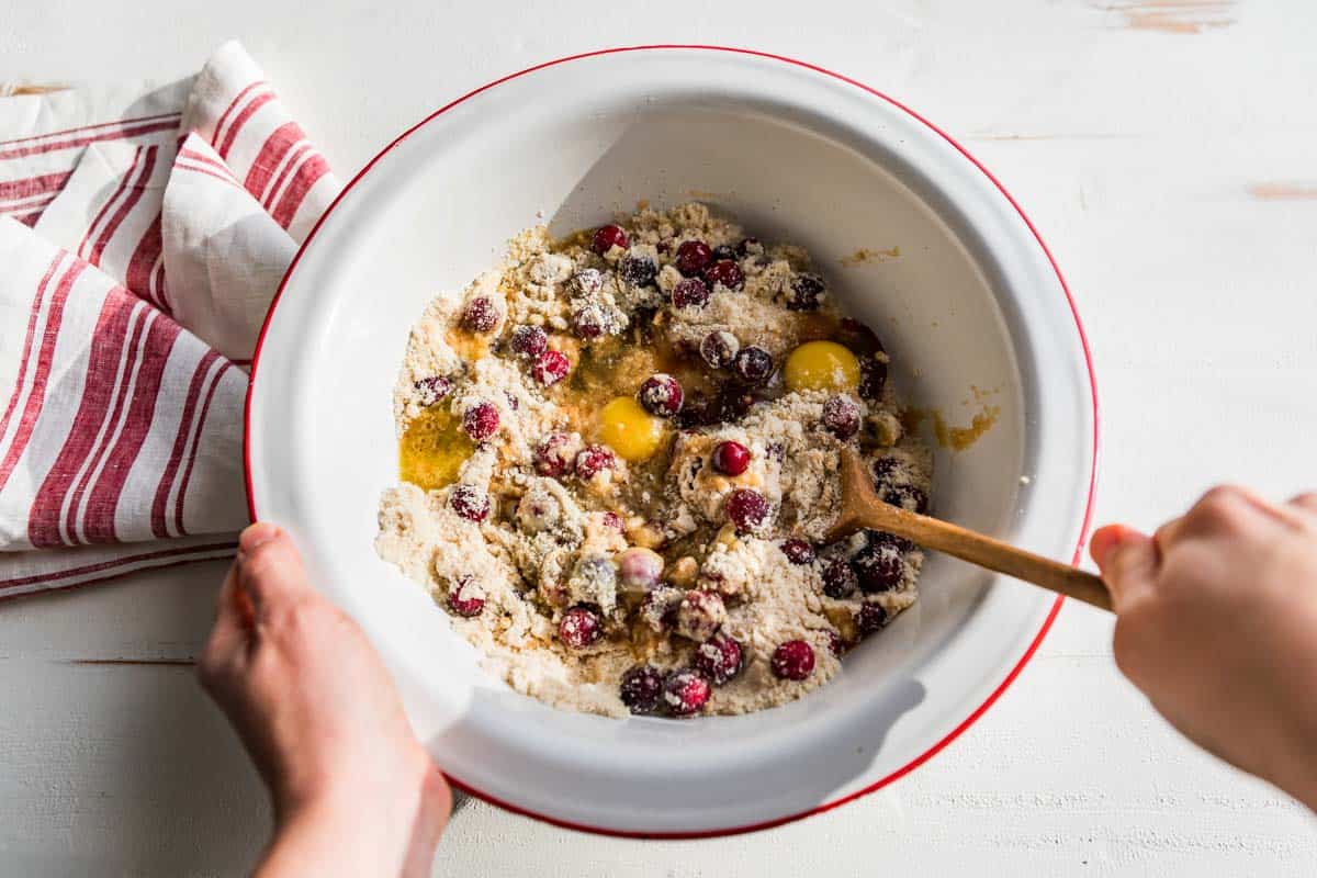 Mixing the cranberries, eggs, vanilla, and melted butter into the dry ingredients in a red rimmed white bowl.