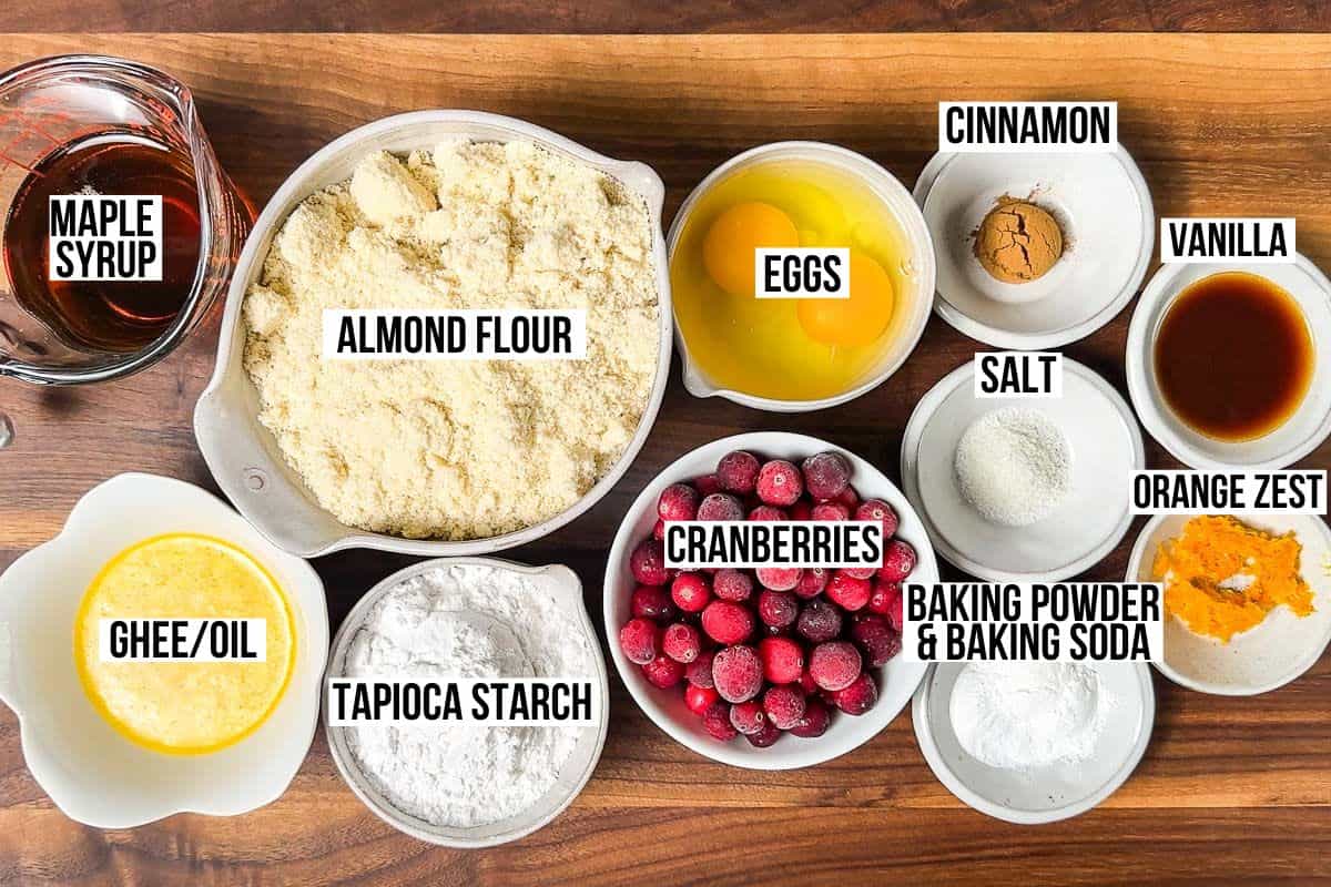 Almond flour, tapioca starch, maple syrup, ghee, cranberries, eggs, vanilla, cinnamon, orange zest, and baking powder in dishes on a wood cutting board.