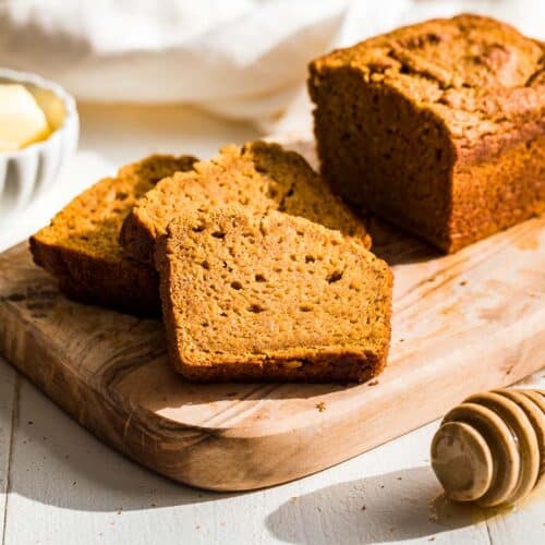 A loaf of paleo pumpkin bread with three slices cut out on a wood cutting board with butter and honey in the background.