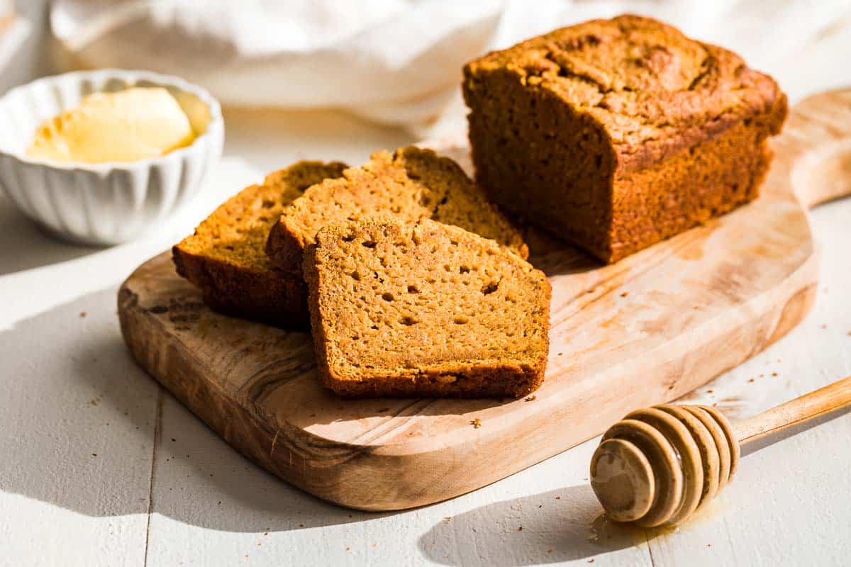 A loaf of paleo pumpkin bread with three slices cut out on a wood cutting board with butter and honey in the background.