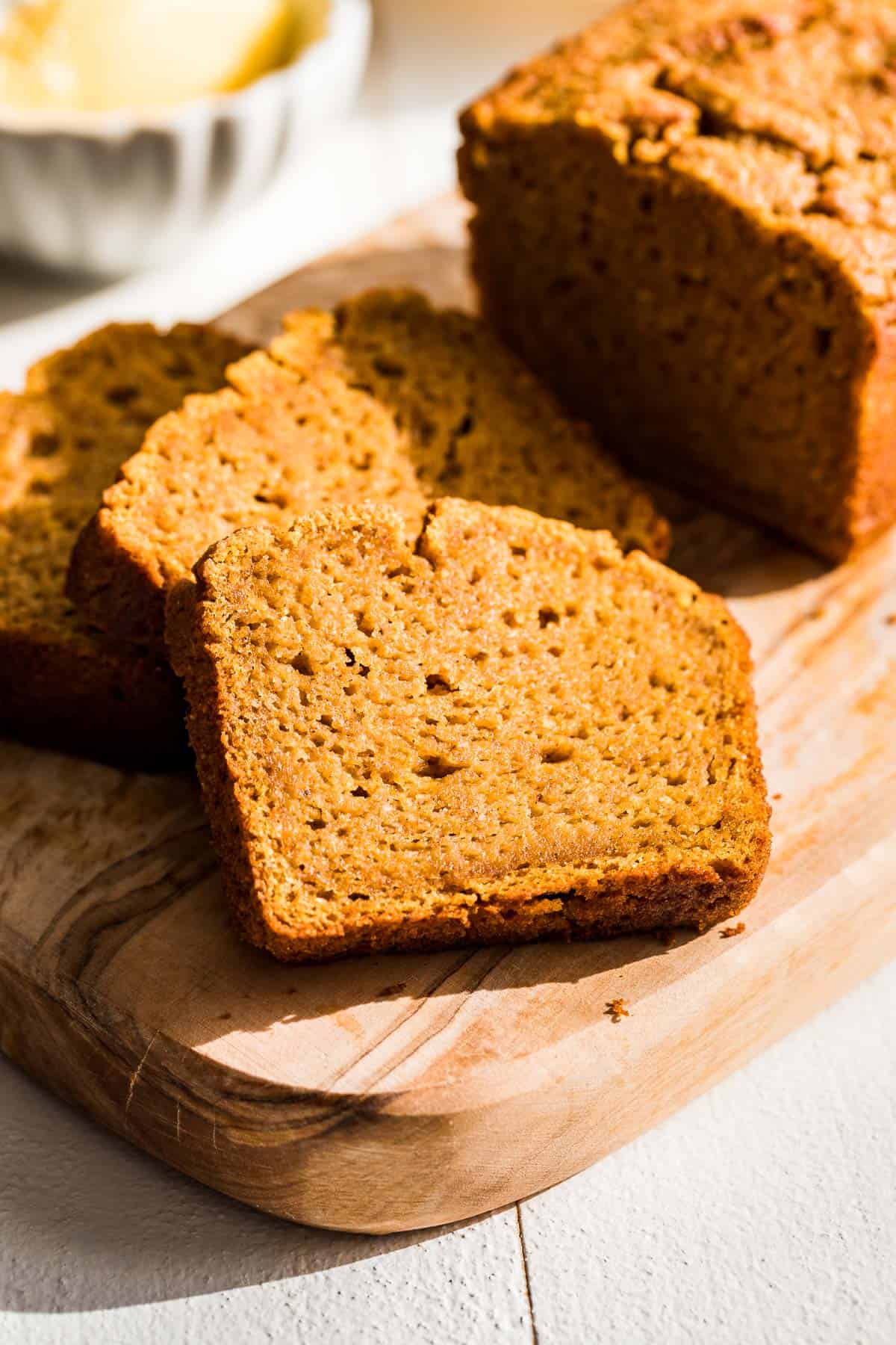 Slices of Paleo Pumpkin Bread on a wood cutting board with butter in the background.