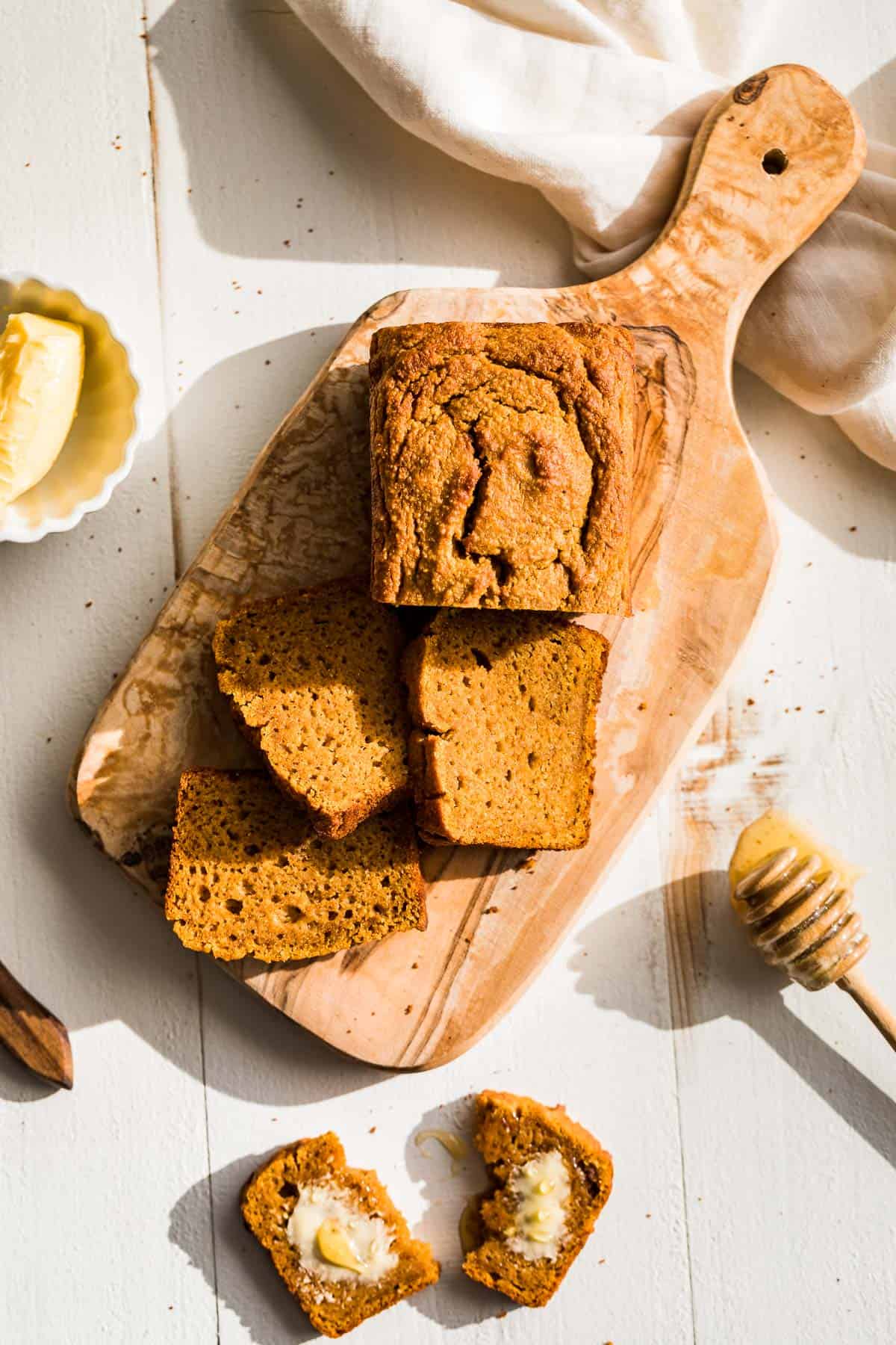 A loaf of paleo pumpkin bread on a wood cutting board with three slices cut and one slice buttered.