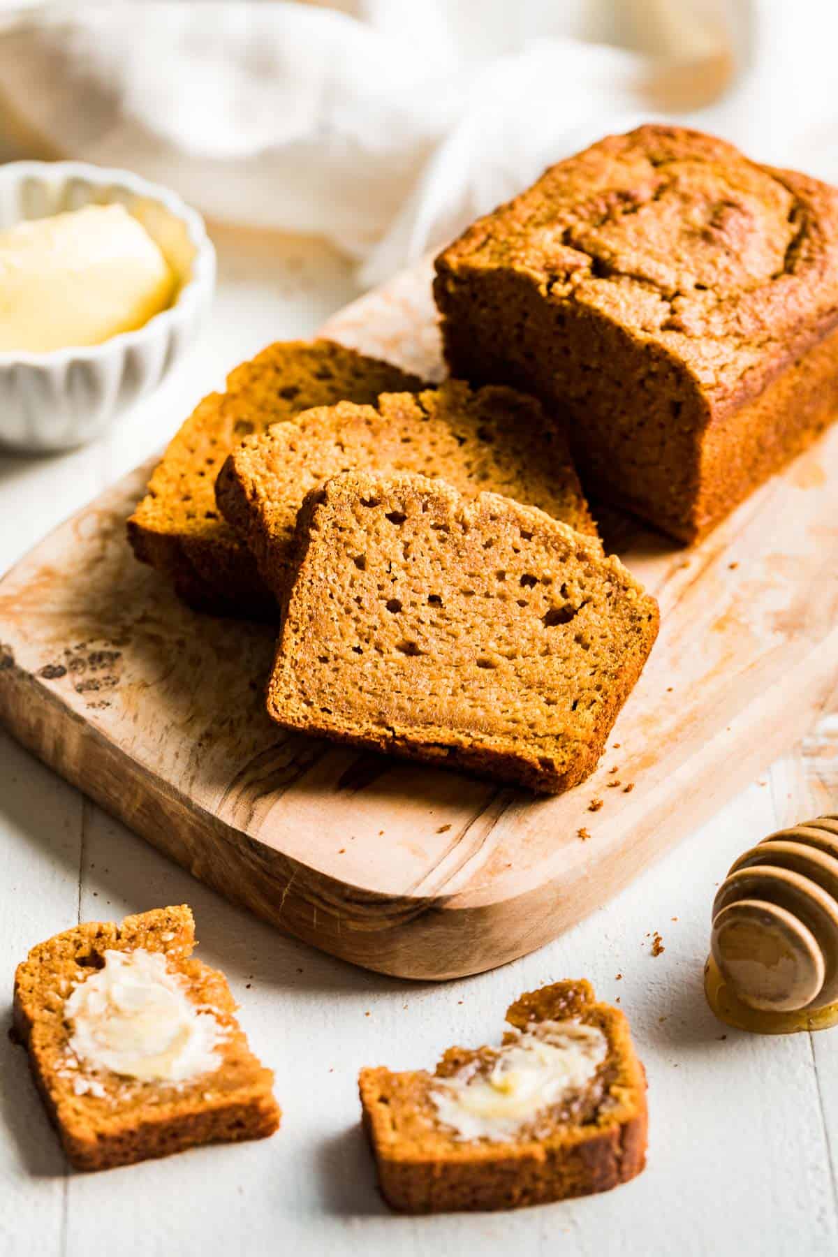 A loaf of Paleo Pumpkin Bread sliced on a wood cutting board with a slice buttered in the front.