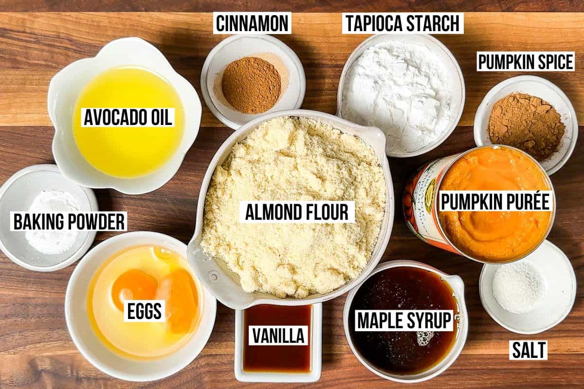Almond flour, tapioca starch, maple syrup, pumpkin purée, pumpkin spice, cinnamon, eggs, vanilla, and baking powder in small containers on a wood cutting board.
