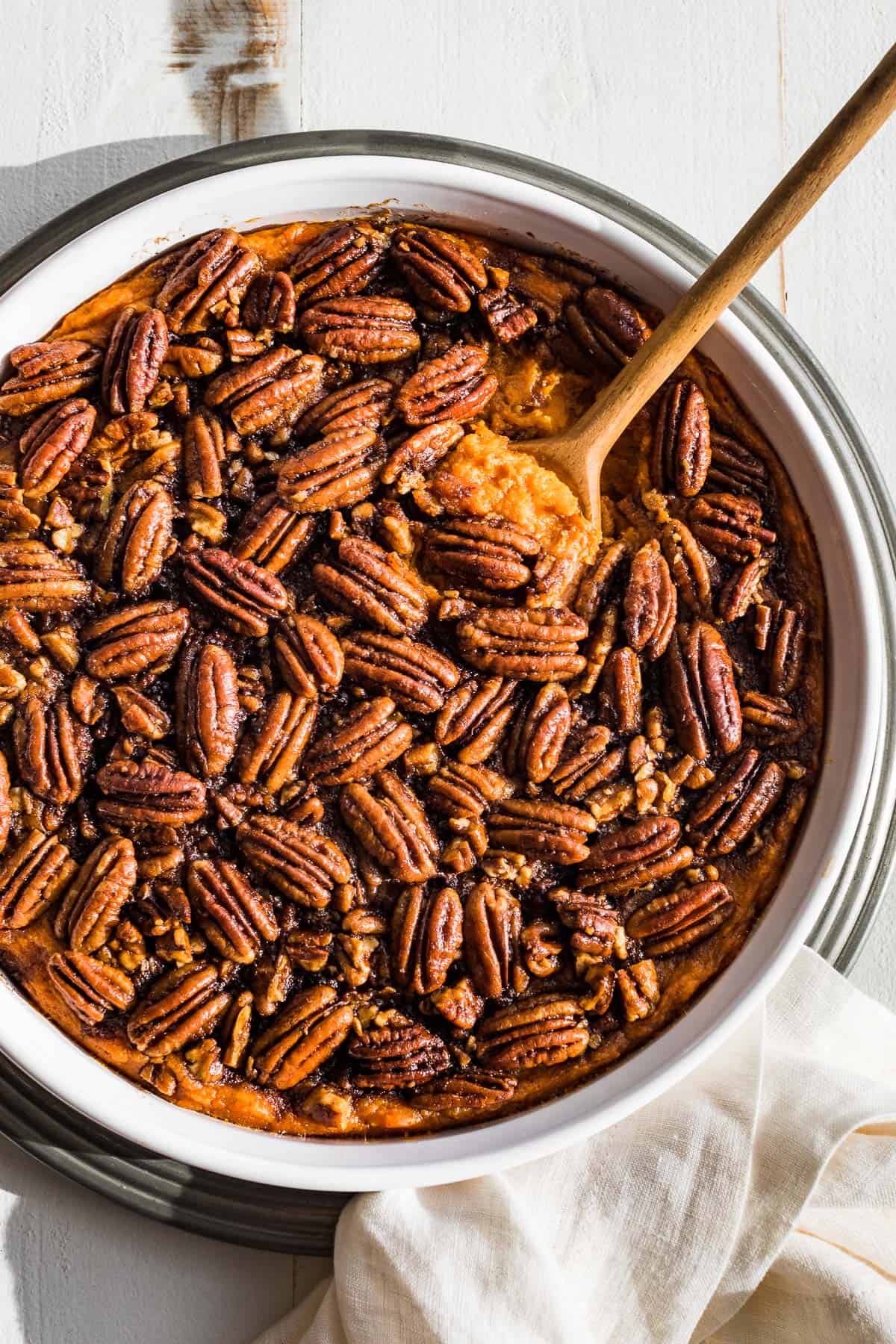 Sweet potato casserole topped with a pecan topping in a white baking dish with a wood spoon scooping out a bite.