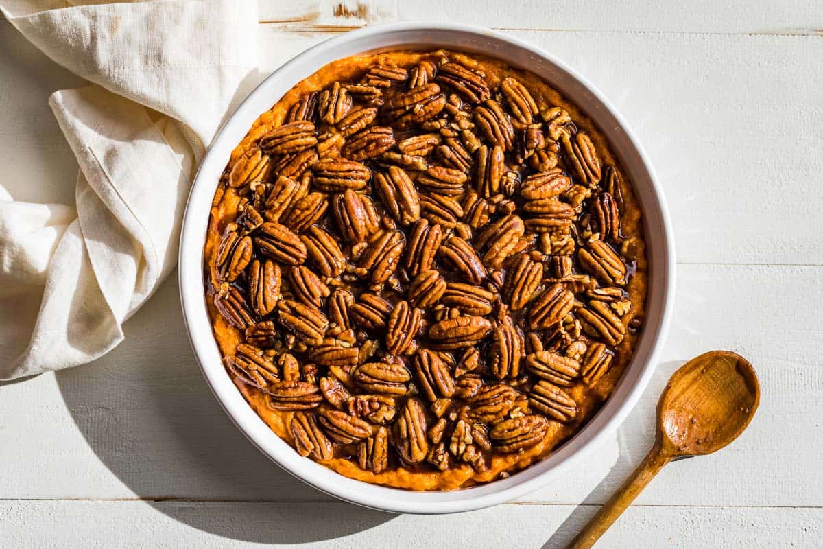 Adding the pecan topping over the top of the sweet potato casserole in a round white baking dish.