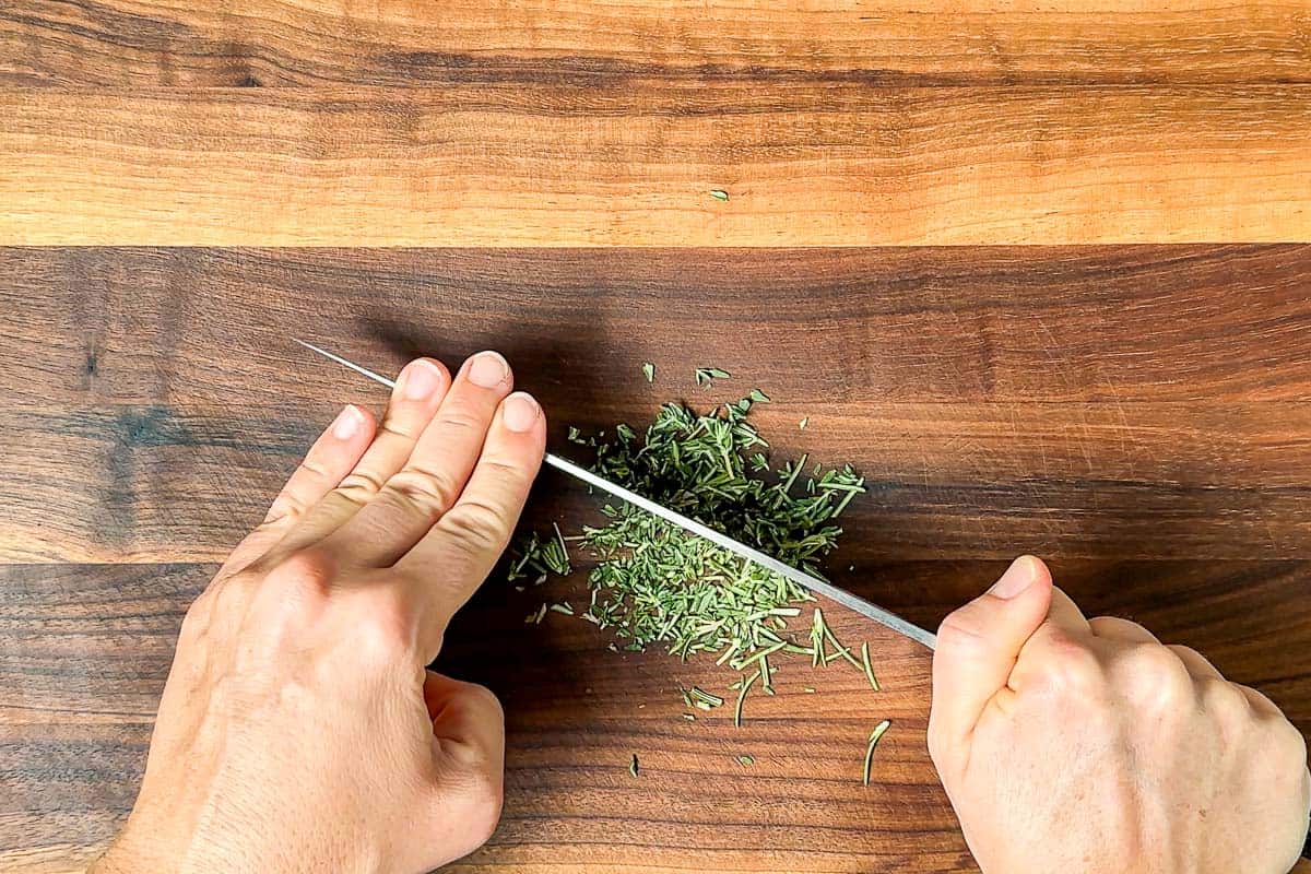 Chopping the thyme and rosemary on a wood cutting board with a chefs knife.