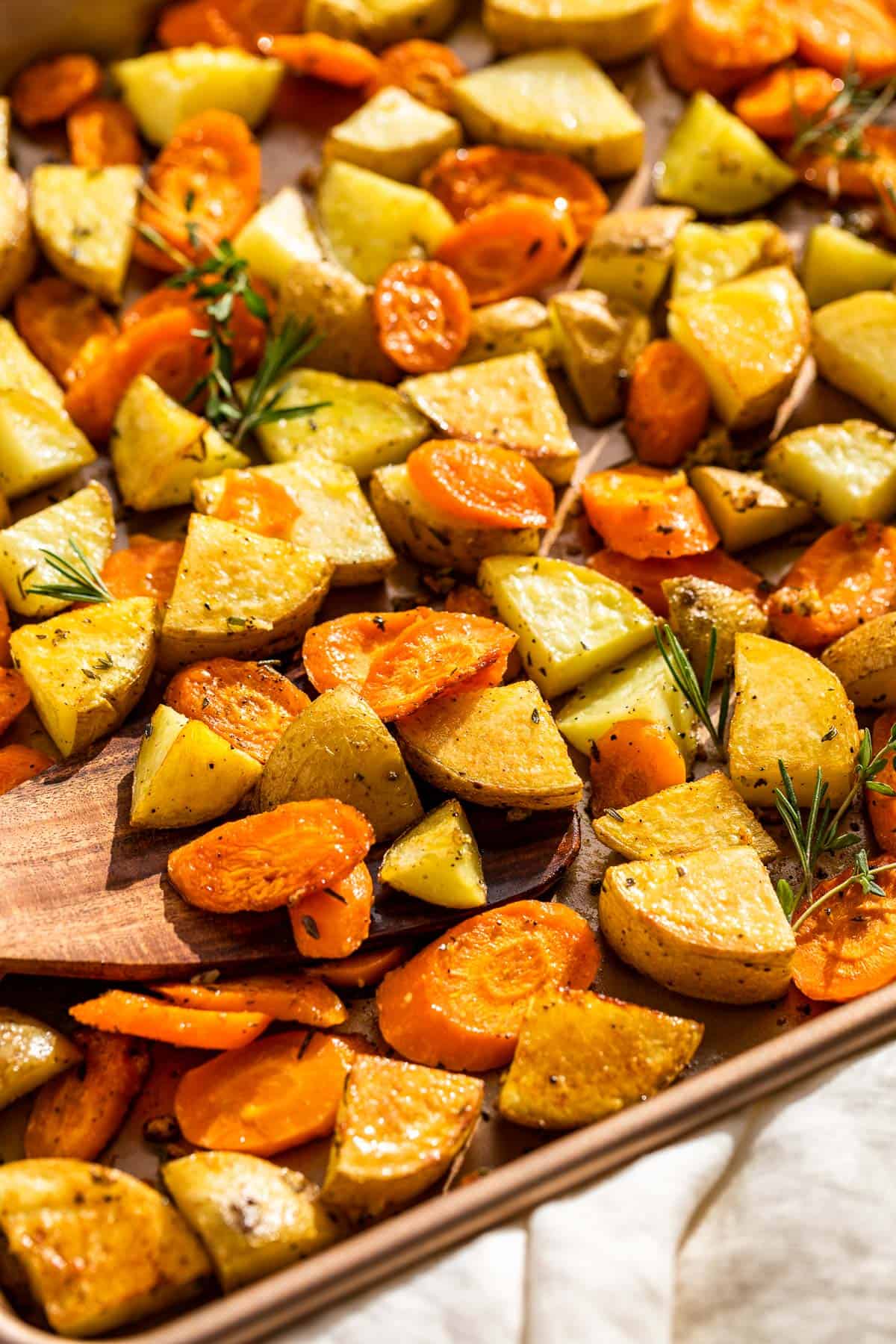 Roasted potatoes and carrots on a baking sheet with a wood spatula scooping some up.