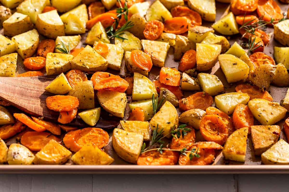 Finished roasted potatoes and carrots on a baking sheet with a wood spatula scooping up some.