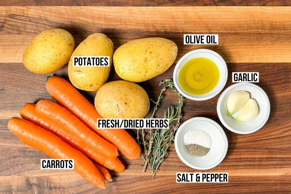 Potatoes, carrots, thyme, rosemary, salt and pepper, garlic, and olive oil in bowls on a wood cutting board.