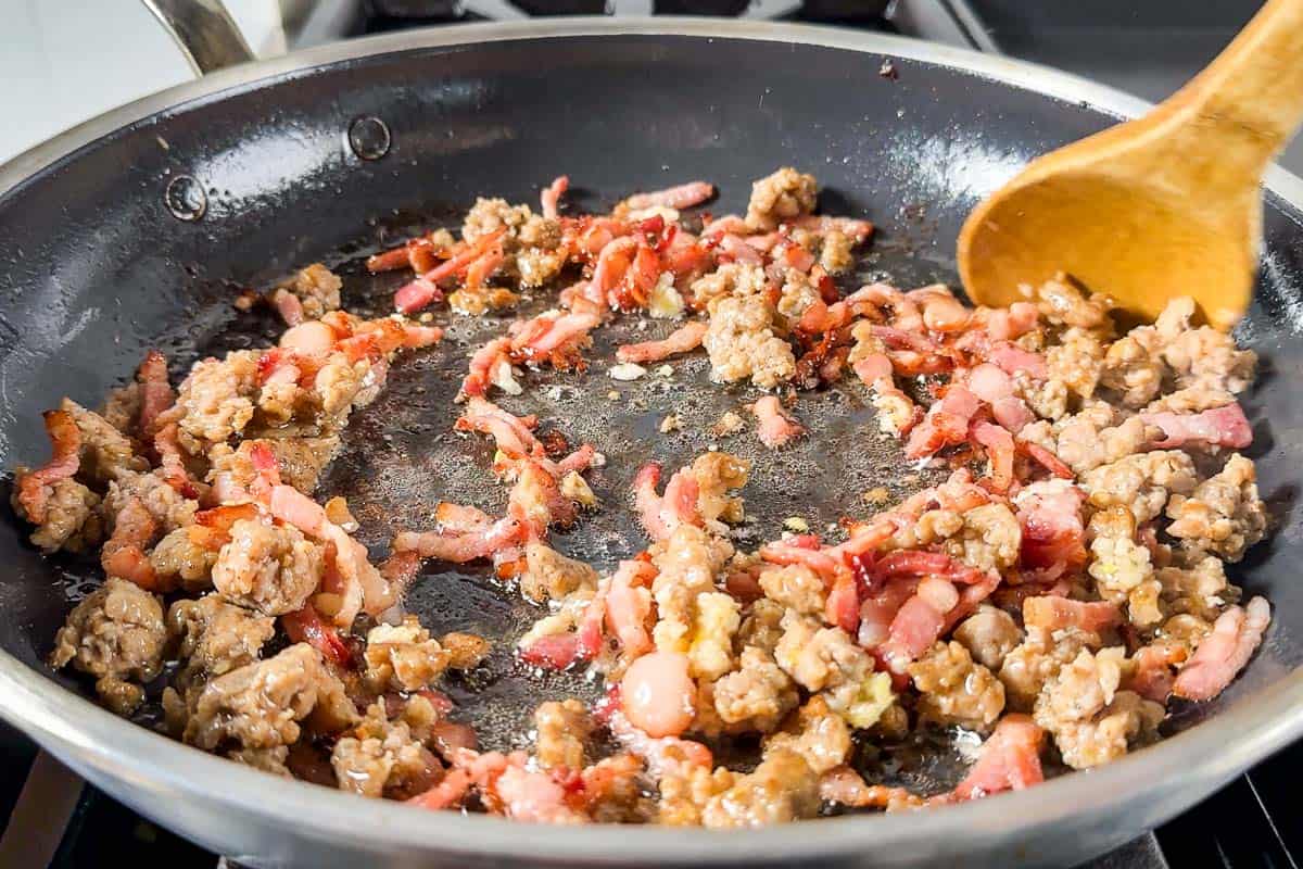 Cooking the ground sausage and diced bacon in a skillet on the stovetop.