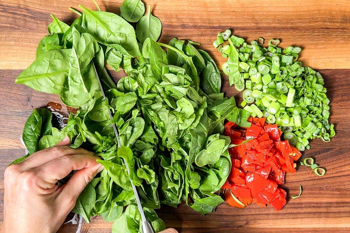 Chopping the spinach, green onions, and roasted red peppers on a wood cutting board with a chefs knife.