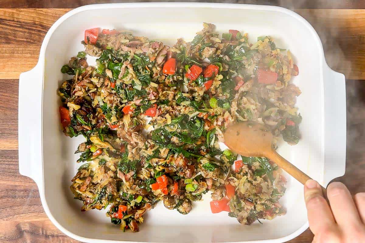 Spreading out the bacon hash brown veggie mixture on the bottom of a large white baking dish.