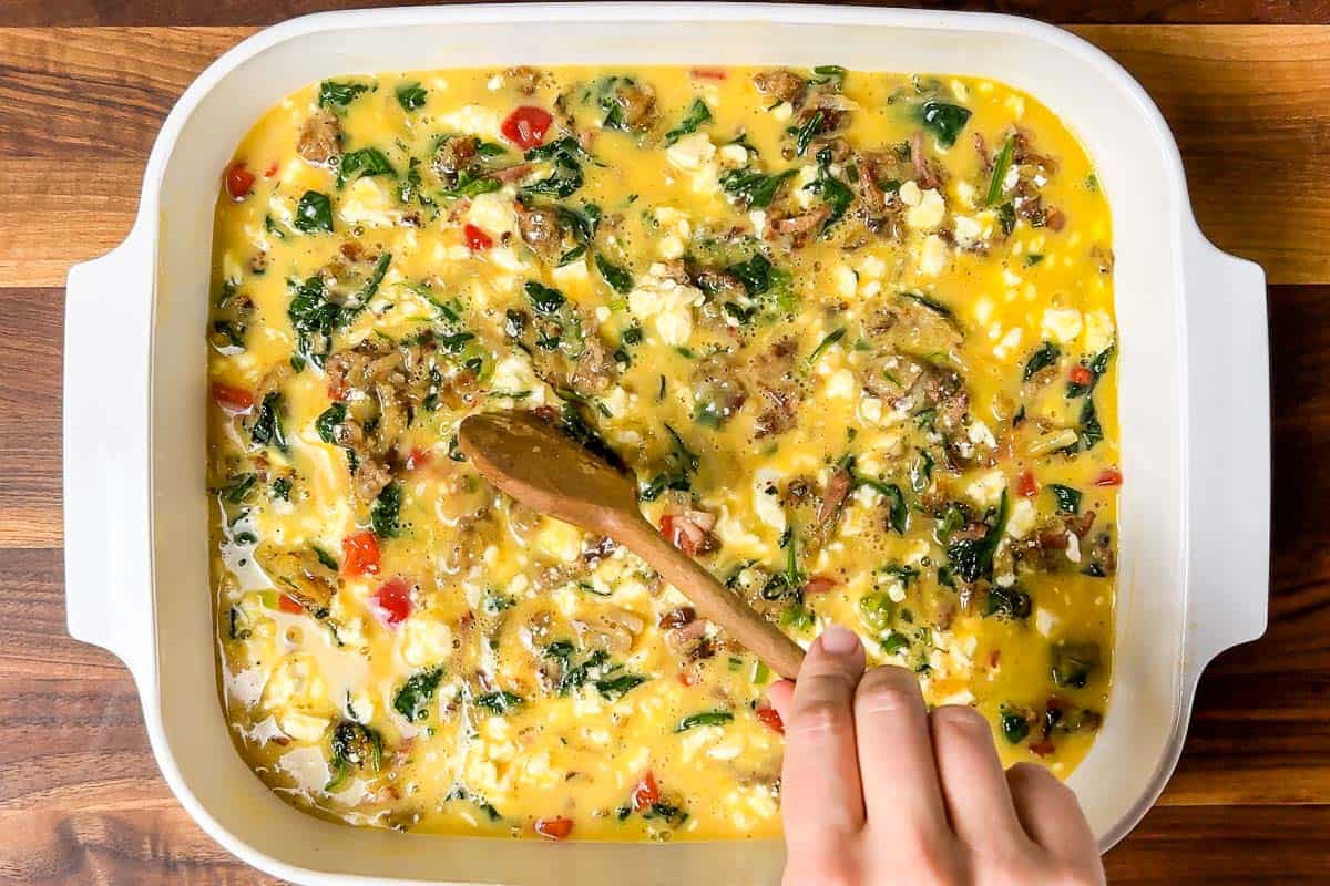 Mixing the bacon hash brown veggie mixture together with the egg mixture in the white baking dish.