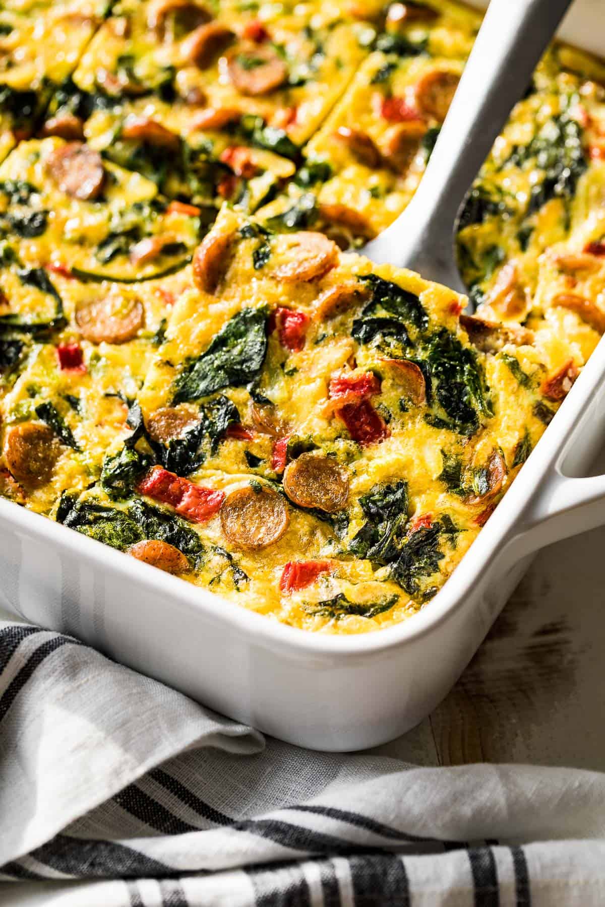 A white baking dish with sliced sausage hash brown breakfast casserole and one slice being lifted out.