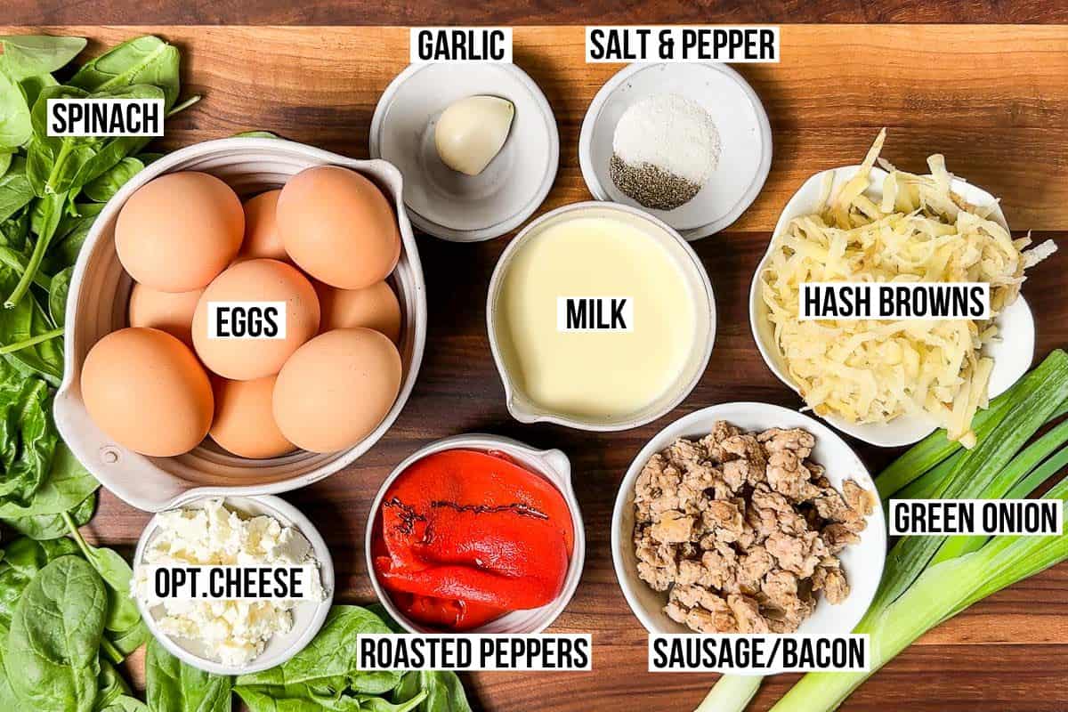 Egg, sausage, garlic, roasted red peppers, hash browns, green onions, salt and pepper, spinach and feta cheese in bowls on a wood cutting board.