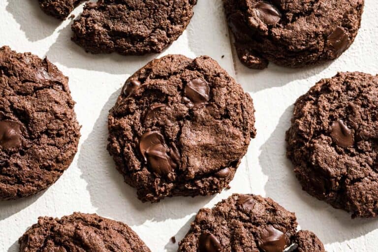 Six chocolate almond butter cookies on a white background with one cookie broken in half.