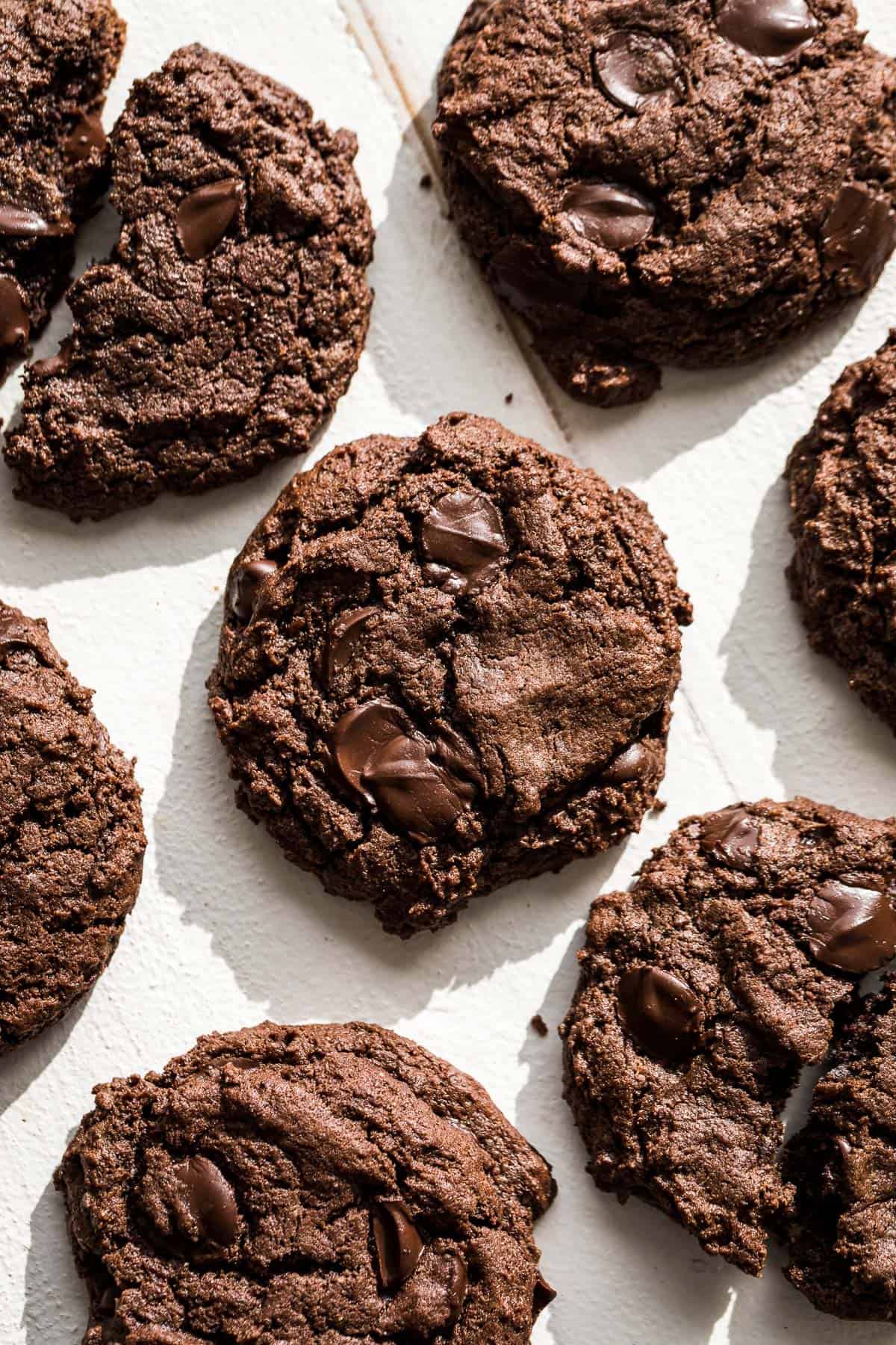 Seven chocolate almond butter cookies on a white background with one cookie broken in half.