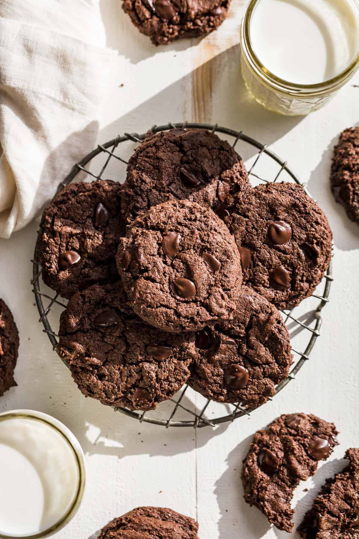 Chocolate almond butter cookies stacked on a round wire cooling rack with two glasses of milk on the side.