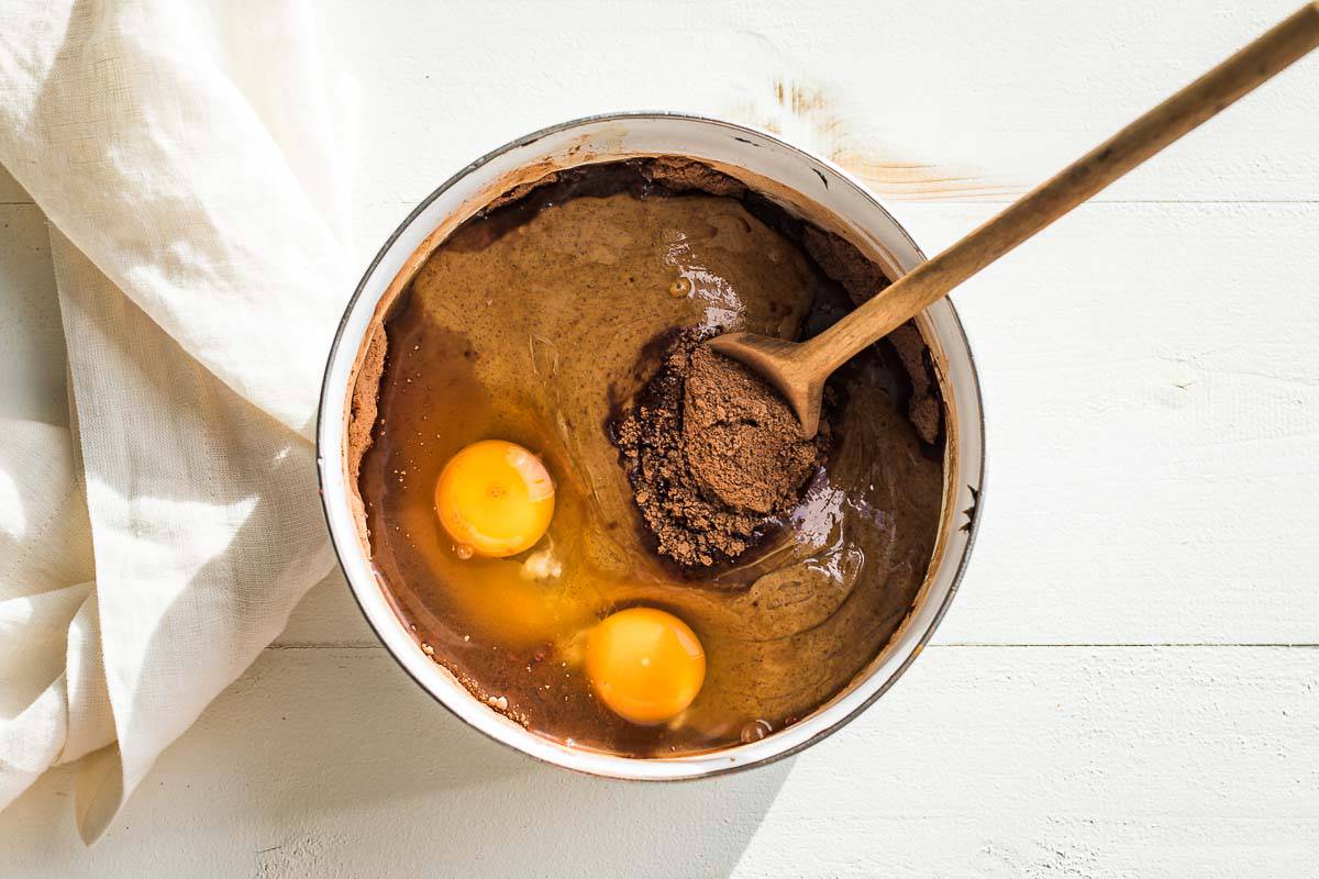 Almond butter, eggs, cocoa powder, and coconut sugar in a white bowl with a wood spoon in it.