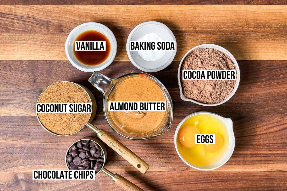 Almond butter, coconut sugar, cocoa powder, eggs, chocolate chips, vanilla, and baking soda in containers on a wood cutting board.