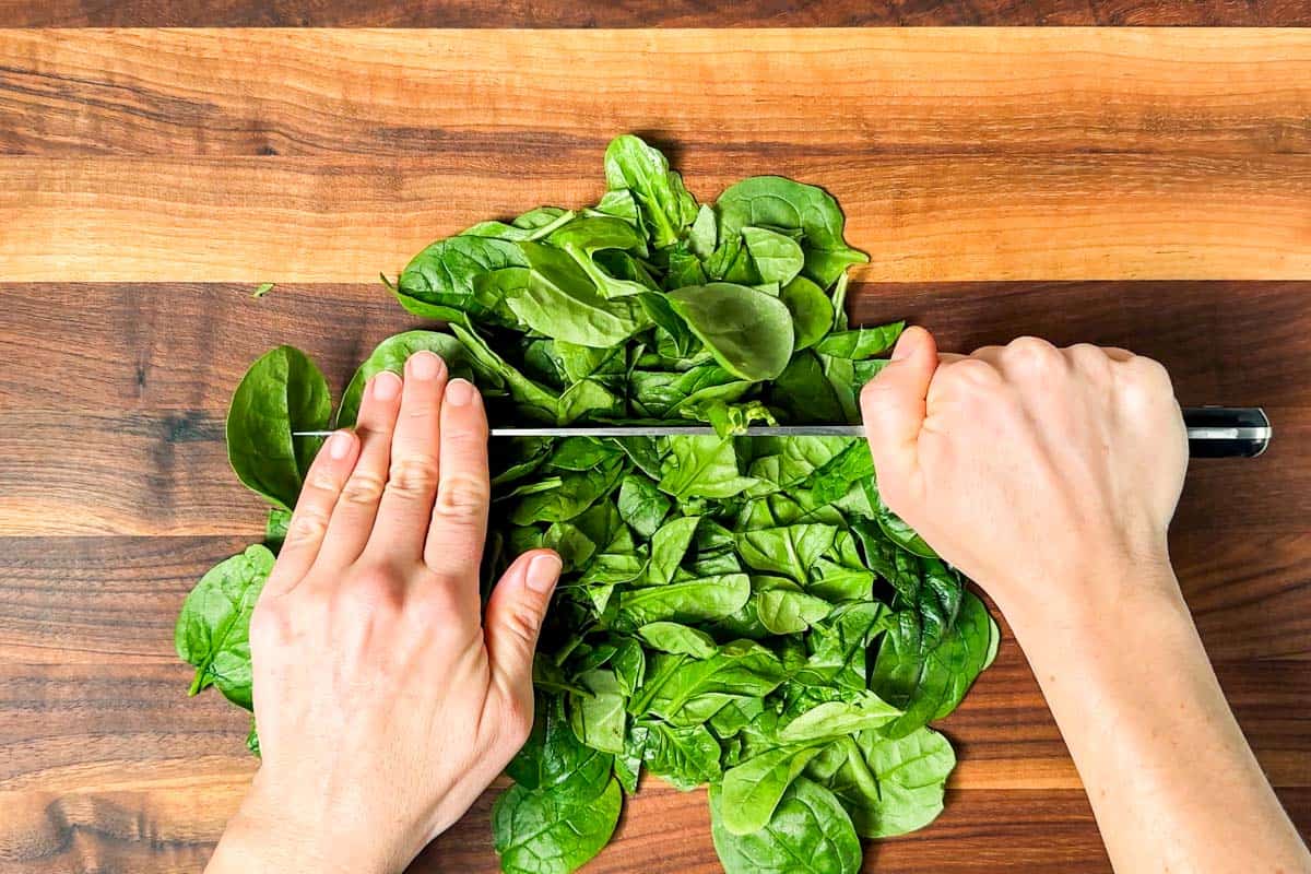 Chopping baby spinach on a wood cutting board with a large chef's knife.