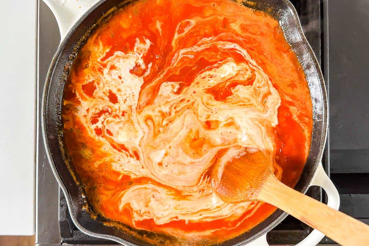 Adding the cream to the tomato sauce in a large white skillet on the stove top.