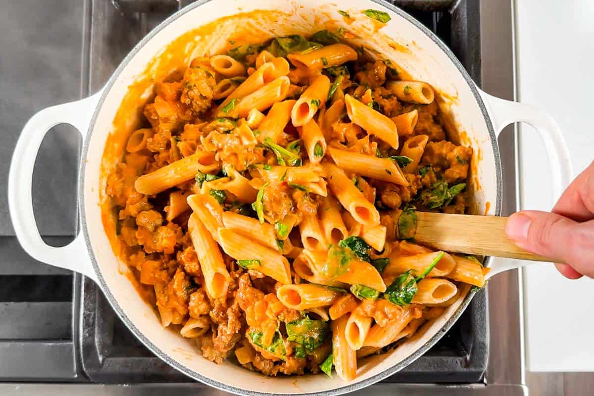 Tossing the tomato cream sauce and chorizo with the cooked pasta in a large white pot with a wood spoon.