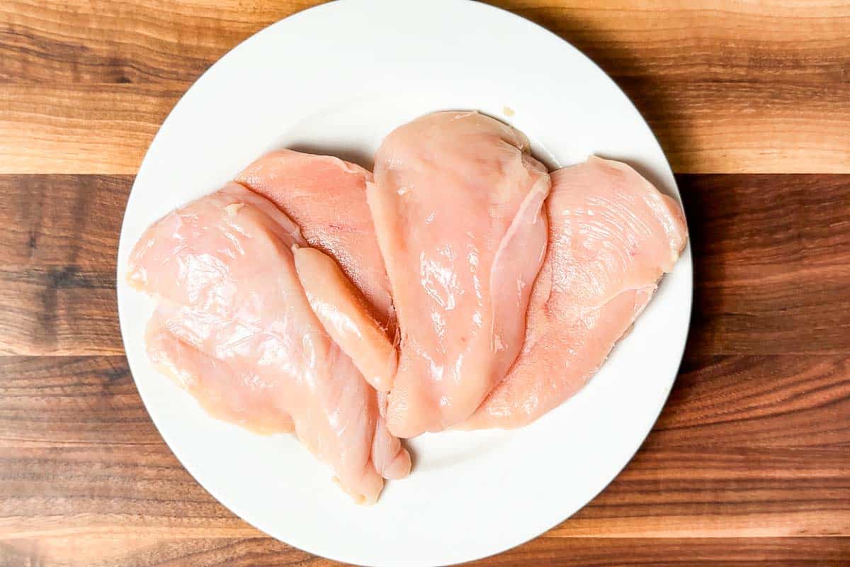 Chicken breasts sliced in half lengthwise on a white plate on a wood cutting board.