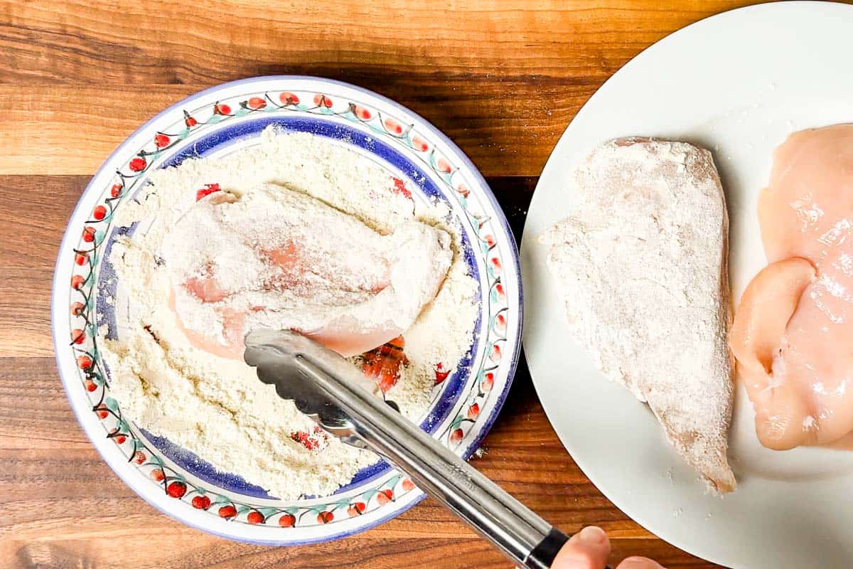 Dredging the chicken breasts in a shallow bowl of flour with metal tongs.