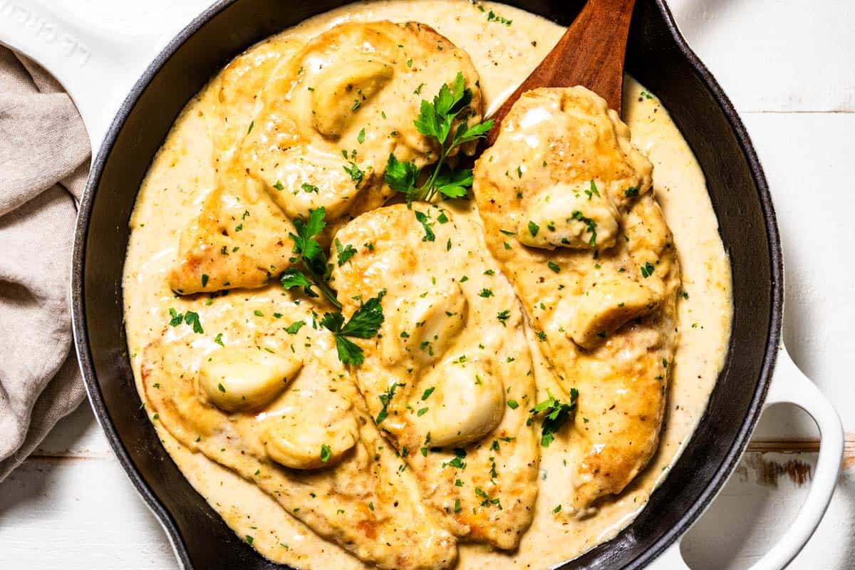 Four chicken breasts topped with garlic cloves in a creamy garlic sauce with a wood spatula on the side of the skillet.