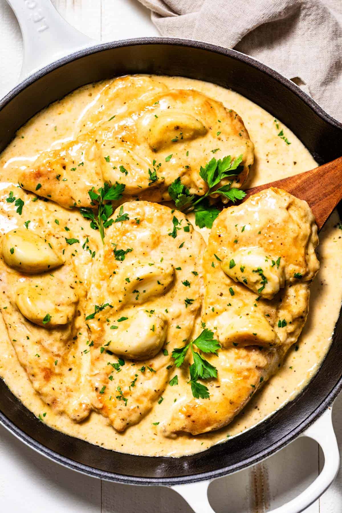 Four chicken breasts in a creamy garlic sauce sprinkled with parsley in a white skillet.