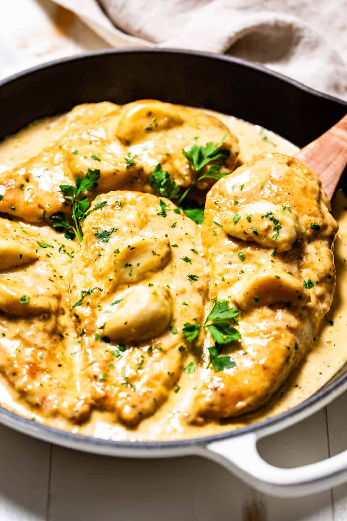 Creamy garlic chicken being lifted out of a white skillet with a wood spoon.