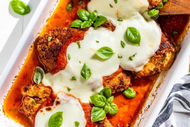 Four pieces of chicken parmesan in a white baking dish topped with melted cheese, red sauce, and sprigs of basil.