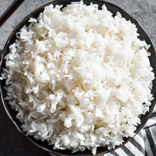 Finished cooked rice in a black bowl with chopsticks on the side and a black and white striped linen on the other side of the bowl.