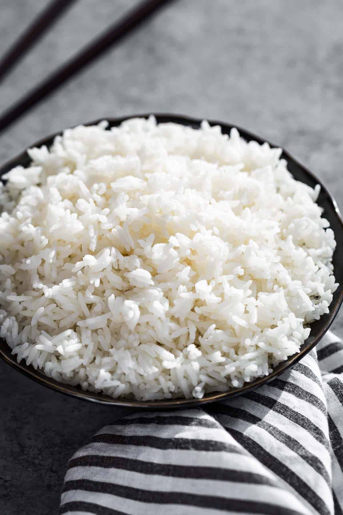 Cooked long grain white rice in a black bowl with chopsticks in the background and a black and white striped linen in the front of the bowl.