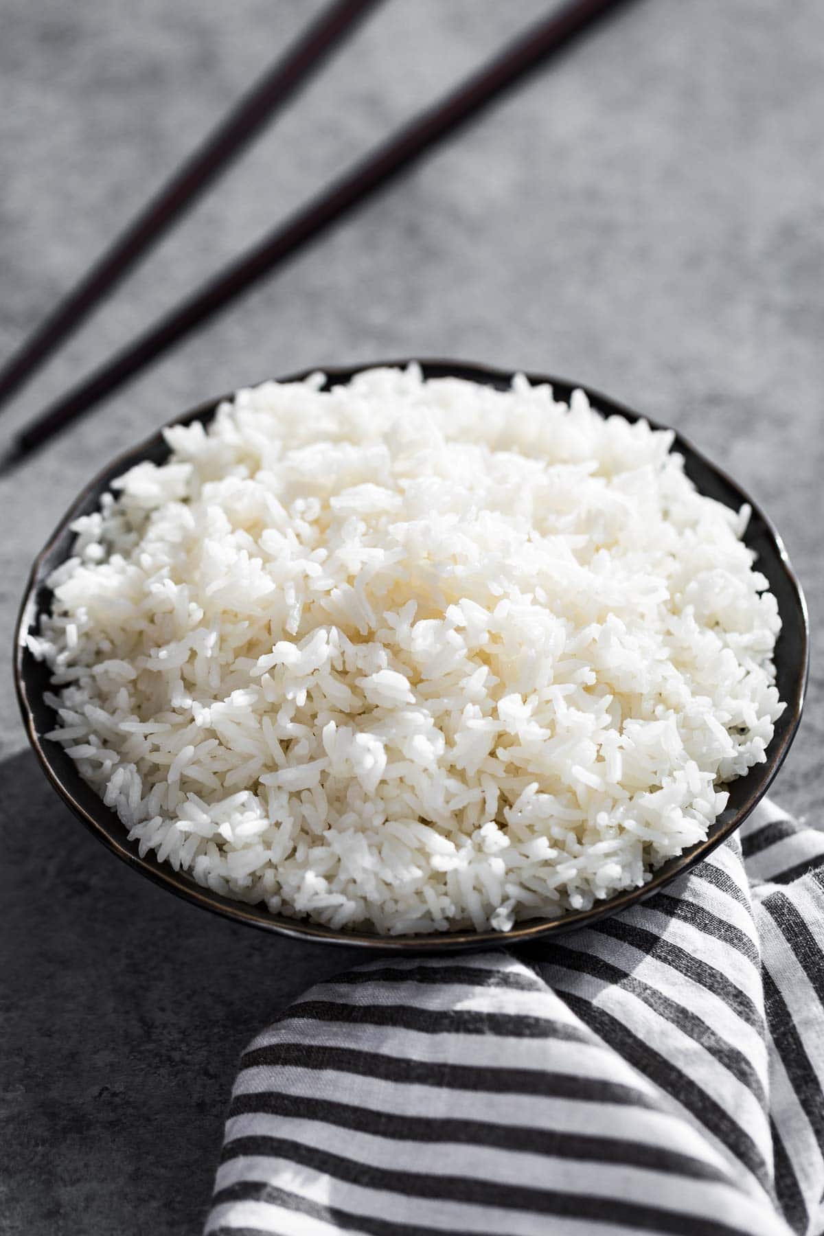 Steamed white rice in a black bowl with chopsticks in the background and black and white striped linen in the front.