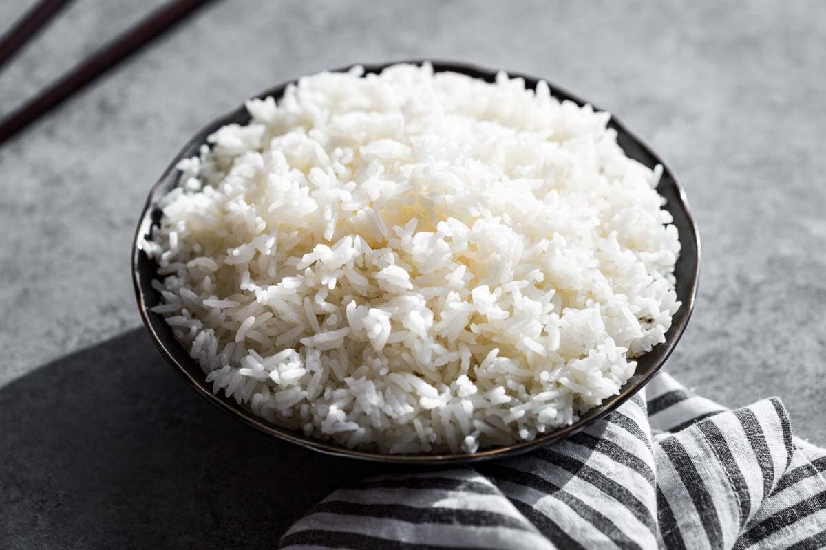 Fluffy cooked white rice in a black bowl on a grey background with chopsticks in the background.