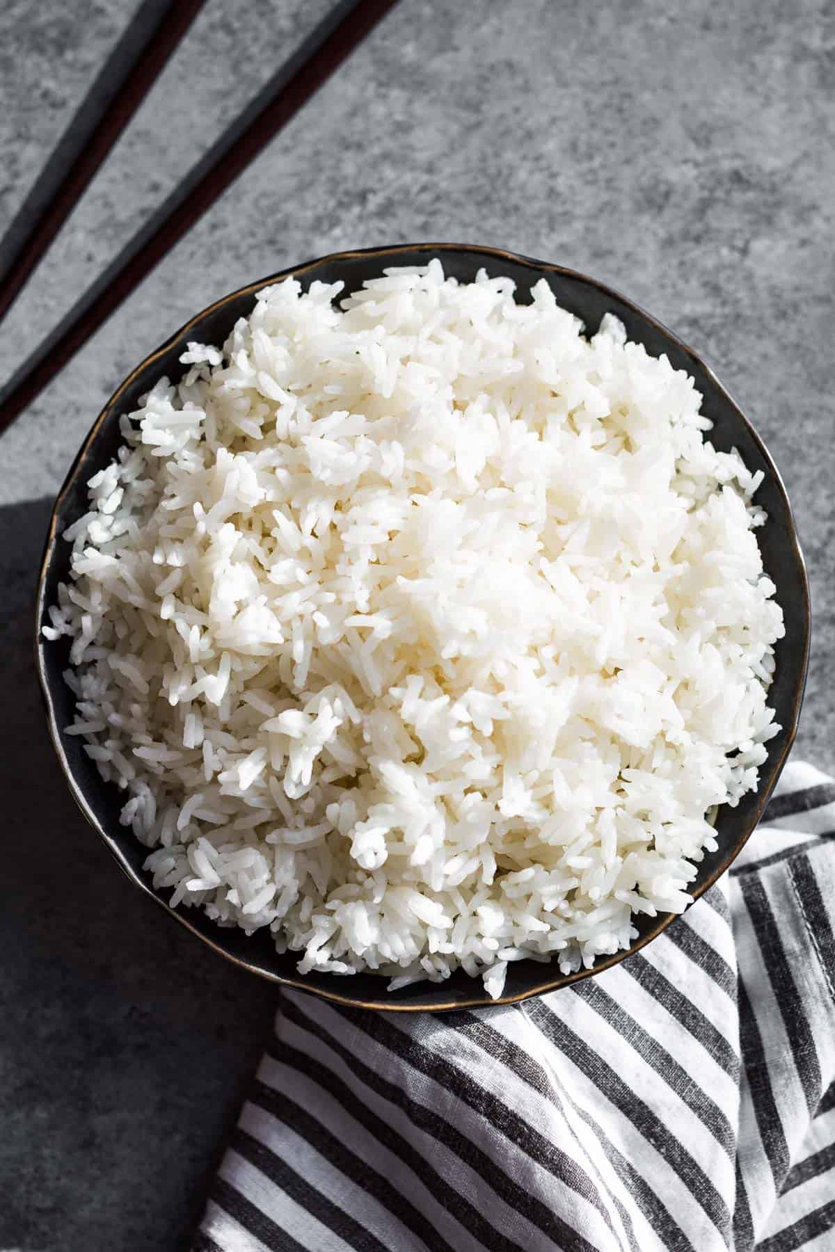 Steamed long grain white rice in a black bowl on a grey background with a black and white striped linen on the side.