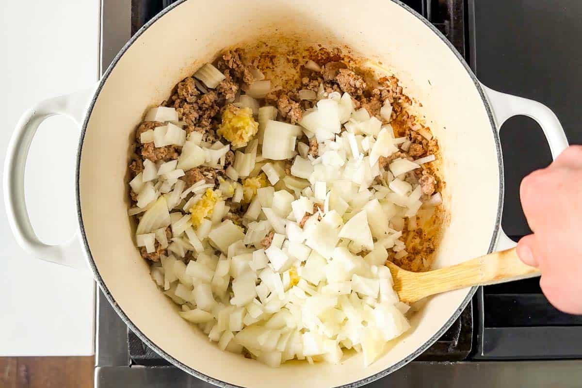 Stirring diced onions, Italian sausage, and garlic with a wood spoon in a large white pot on the stovetop.
