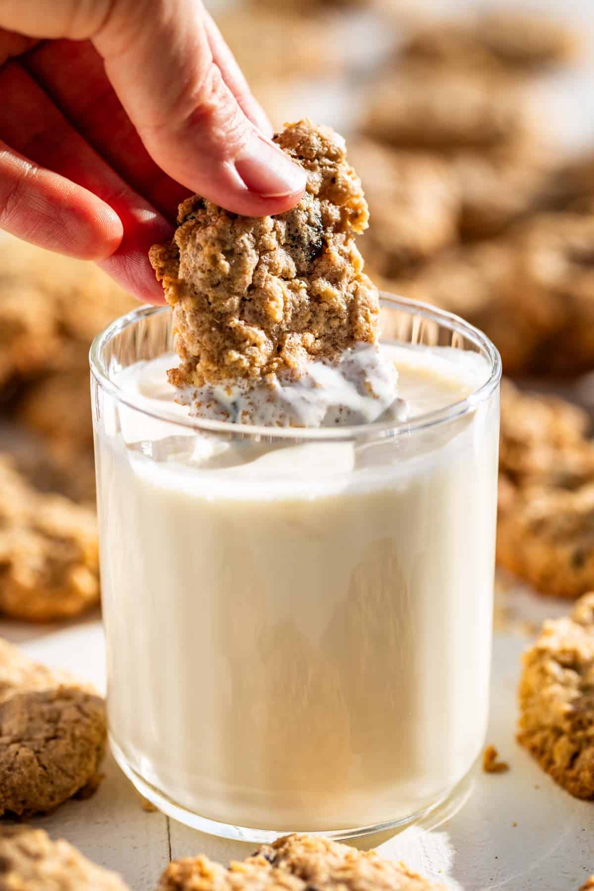 A hand dunking 1/2 of a Oatmeal Raisin Cookie into a glass of milk with cookies around the glass.