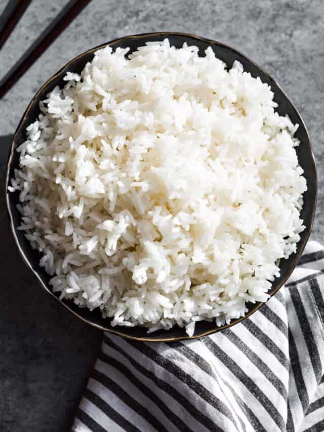 Steamed long grain white rice in a black bowl on a grey background with a black and white striped linen on the side.