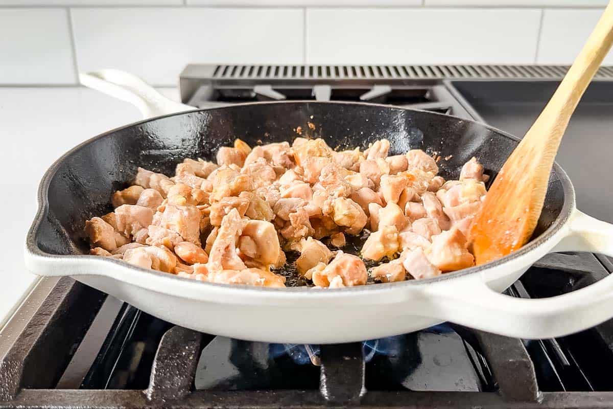 Cooking cubed up chicken thighs in a large white skillet on the stove top with a wooden spoon.