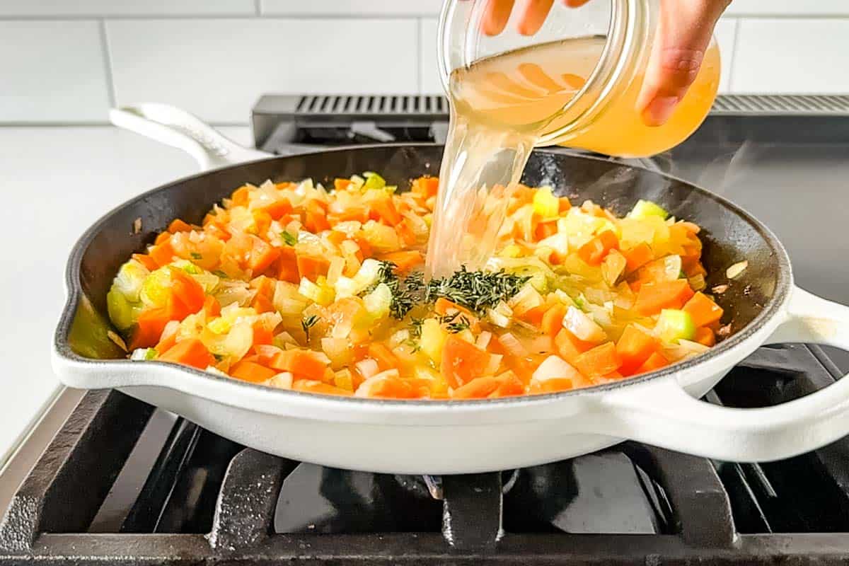 Adding chicken broth and thyme to the cooking diced carrots, onion, and celery in a large white skillet.