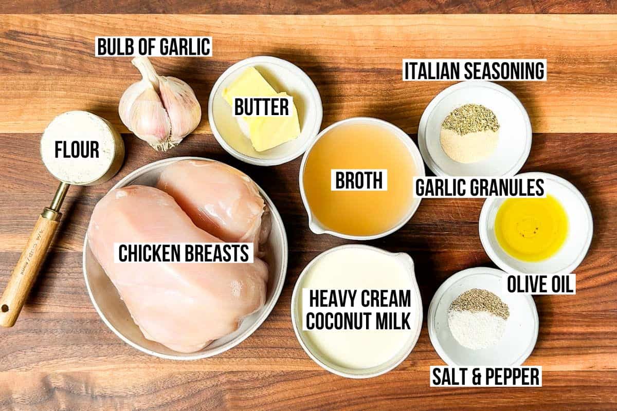 Chicken breasts, flour, a bulb of garlic, butter, broth, creamy, olive oil, garlic powder, Italian seasoning, salt and pepper in bowls on a wood cutting board.