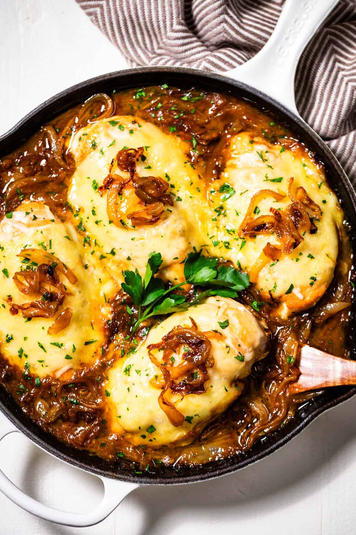 White skillet with four Chicken Breasts topped with cheese sitting in caramelized onions and topped with parsley.