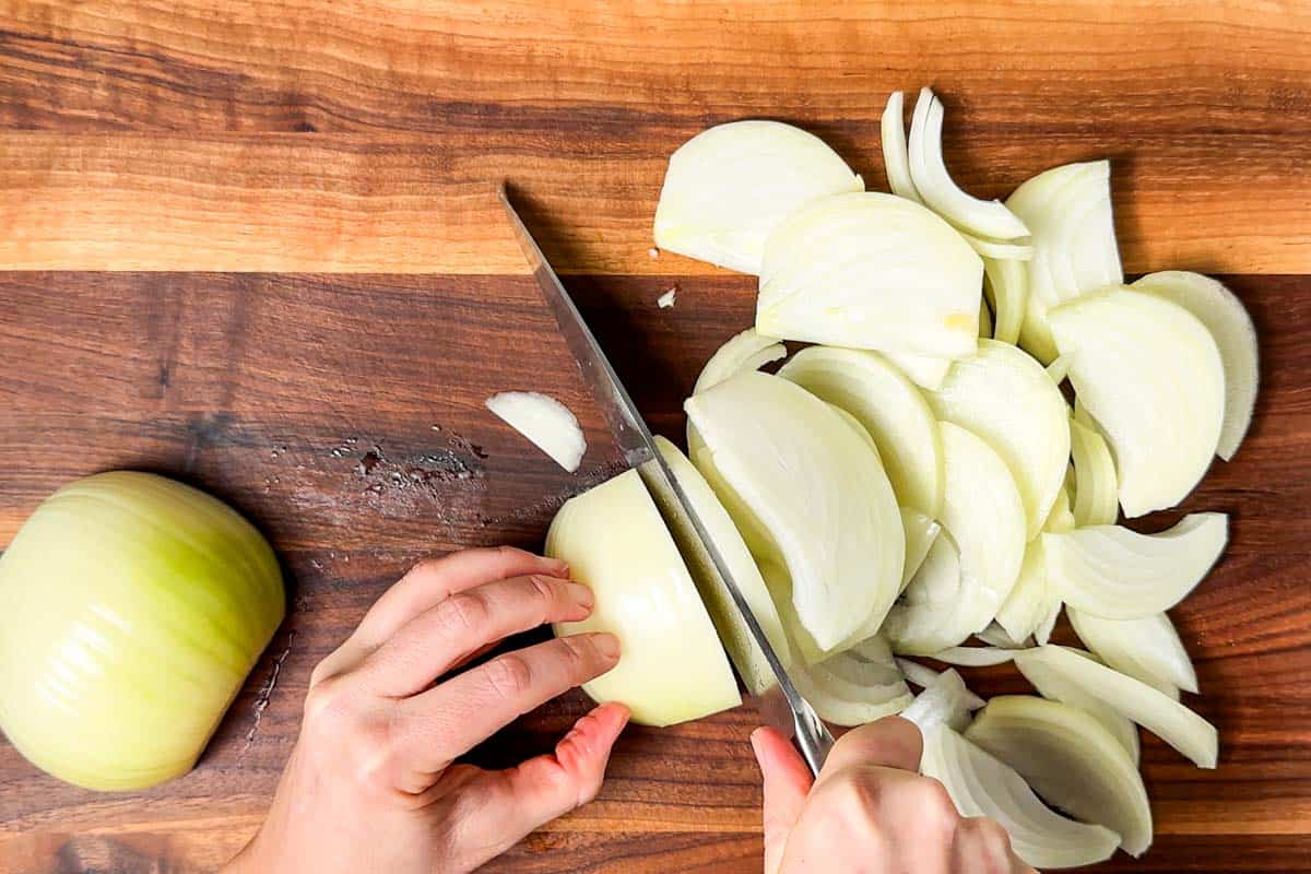 Cutting the onions into half-moon shaped slices with a chef's knife on a wood cutting board.